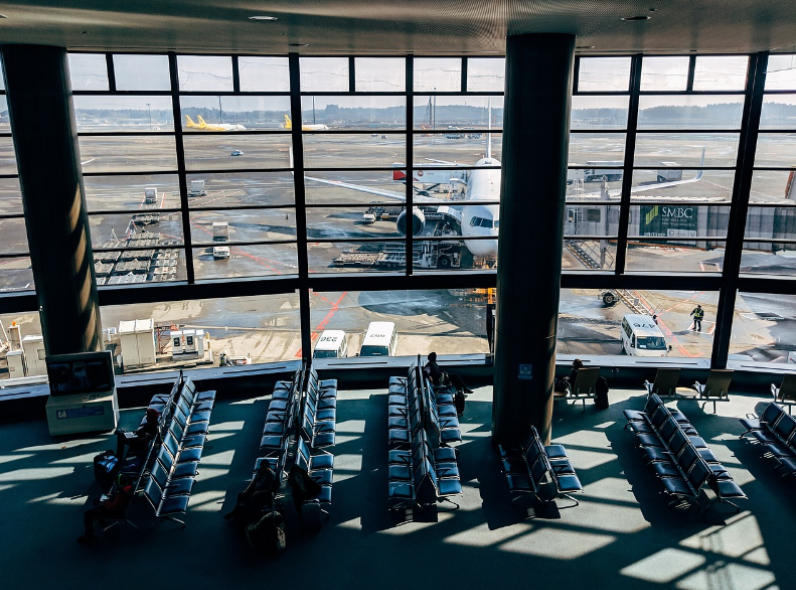 Empty chairs in an airport waiting area overlook a large window with a plane parked at the gate and airport vehicles outside on the tarmac.