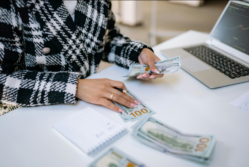 A woman counts cash on a table beside a laptop, focused on her task in a well-lit workspace.