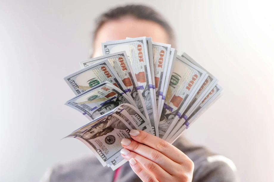 A woman covers her face with a handful of money, emphasizing her expression of surprise or excitement.
